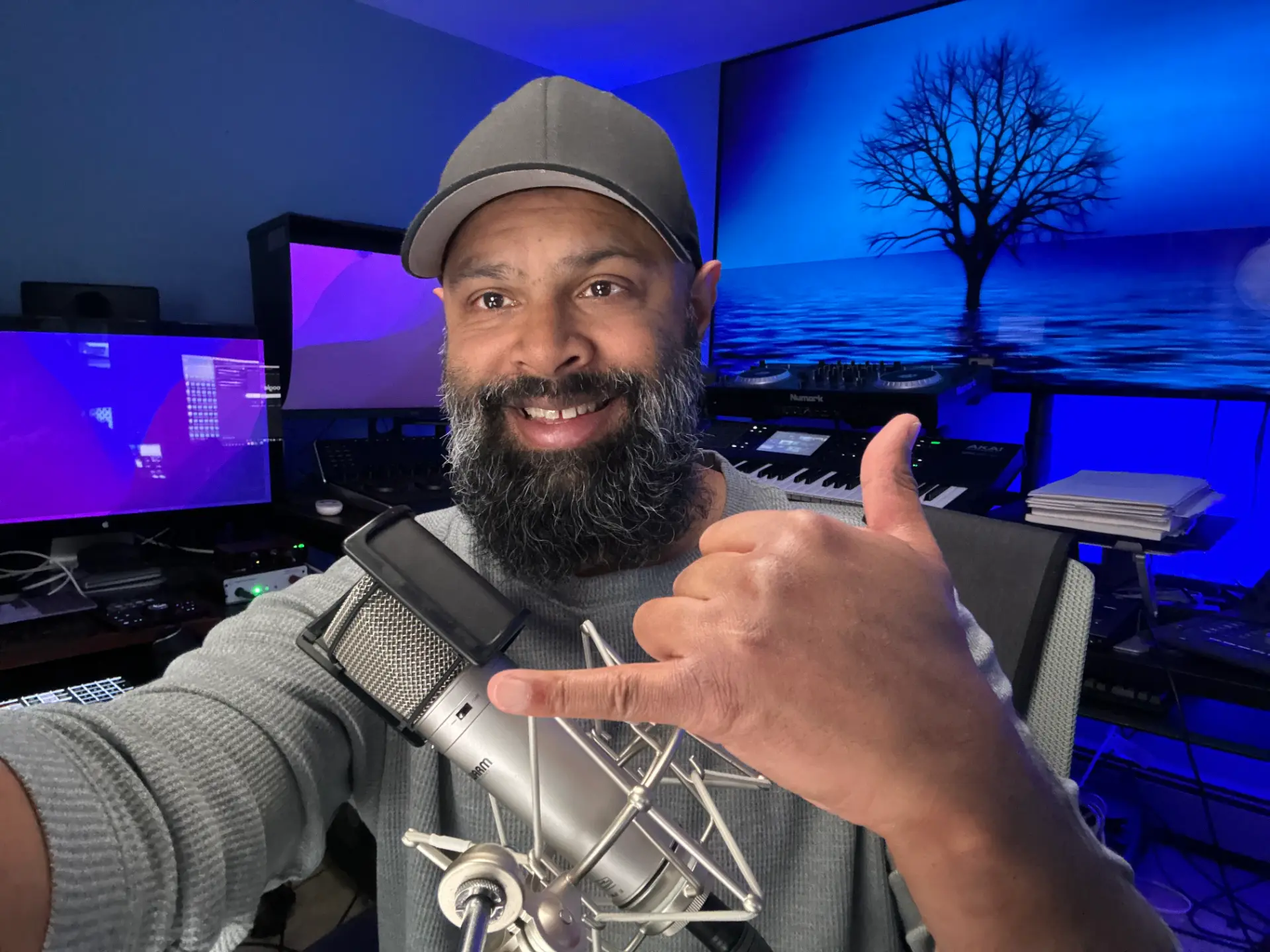 Dee Kumar smiling in his studio behind a microphone, giving a friendly hang-loose sign